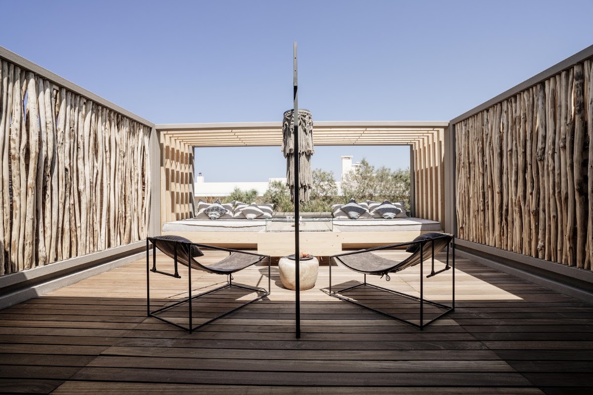 Modern pergola with two lounge chairs and hanging lamp on wooden deck overlooking resort grounds and sea.