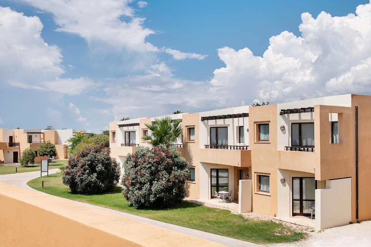 Tan and white resort buildings with balconies and pergolas line a manicured lawn with flowering shrubs under a bright blue sky at VOI Arenella Resort.