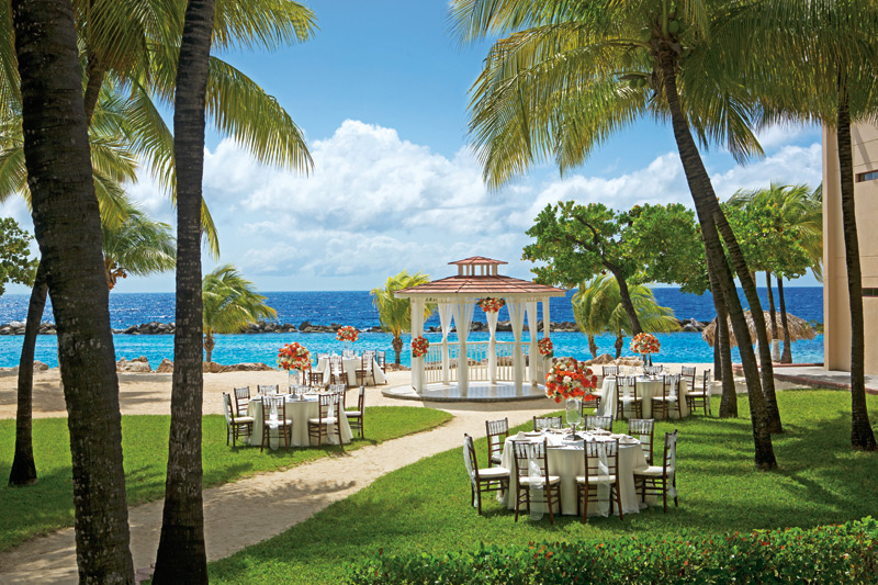 Sunscape Curaçao beachfront ceremony setup with round tables, gazebo, and turquoise water framed by palm trees.