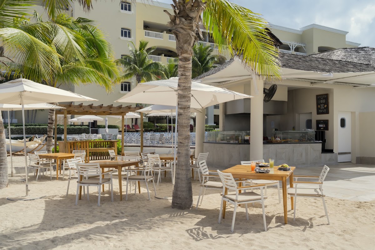 Beachfront dining area at JOIA Rose Hall by Iberostar with wooden tables, white chairs, cream umbrellas, and a tall palm tree, with the resort building and bar in the background.