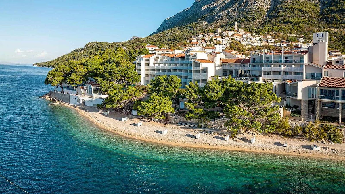 TUI BLUE Makarska resort buildings overlook a pebbly beach and turquoise Adriatic waters, with forested mountains rising behind the coastal town.