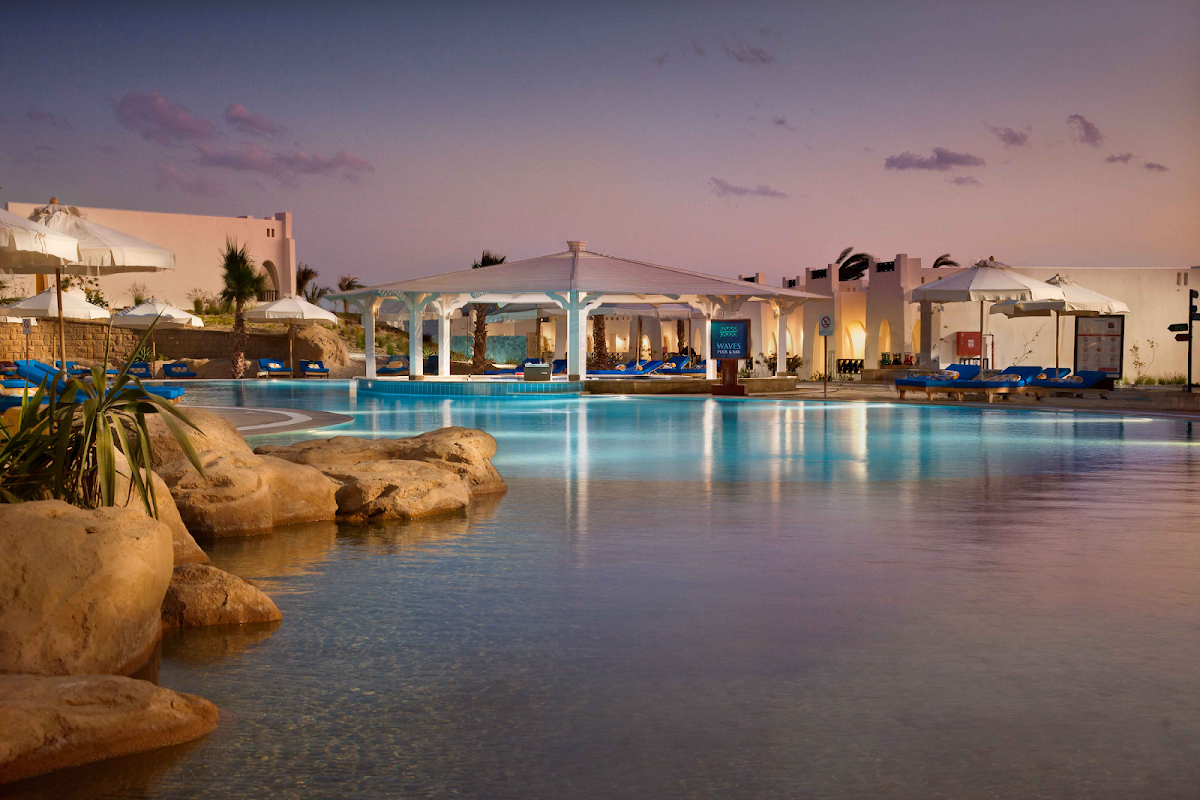 Large illuminated pool with white pergola structures at dusk, surrounded by pale stone buildings and blue loungers at Hilton Marsa Alam Nubian Resort.