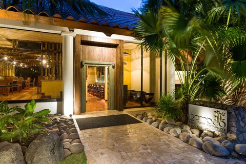 Illuminated beachfront dining pavilion with white railings and thatched-roof bungalows reflected in the pool at dusk.