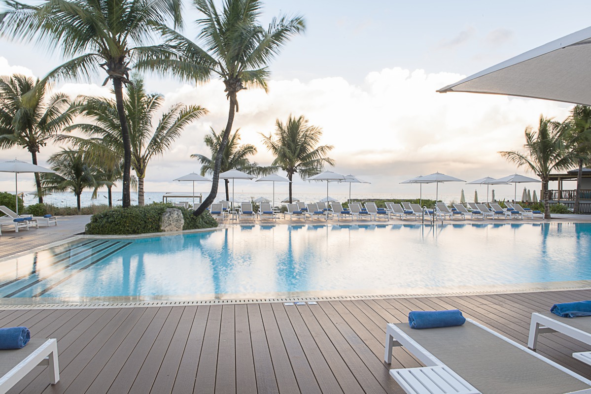 Large curved pool with blue water and wooden deck overlooking the ocean, flanked by palm trees, lounge chairs, and umbrellas at Club Med Columbus Isle.
