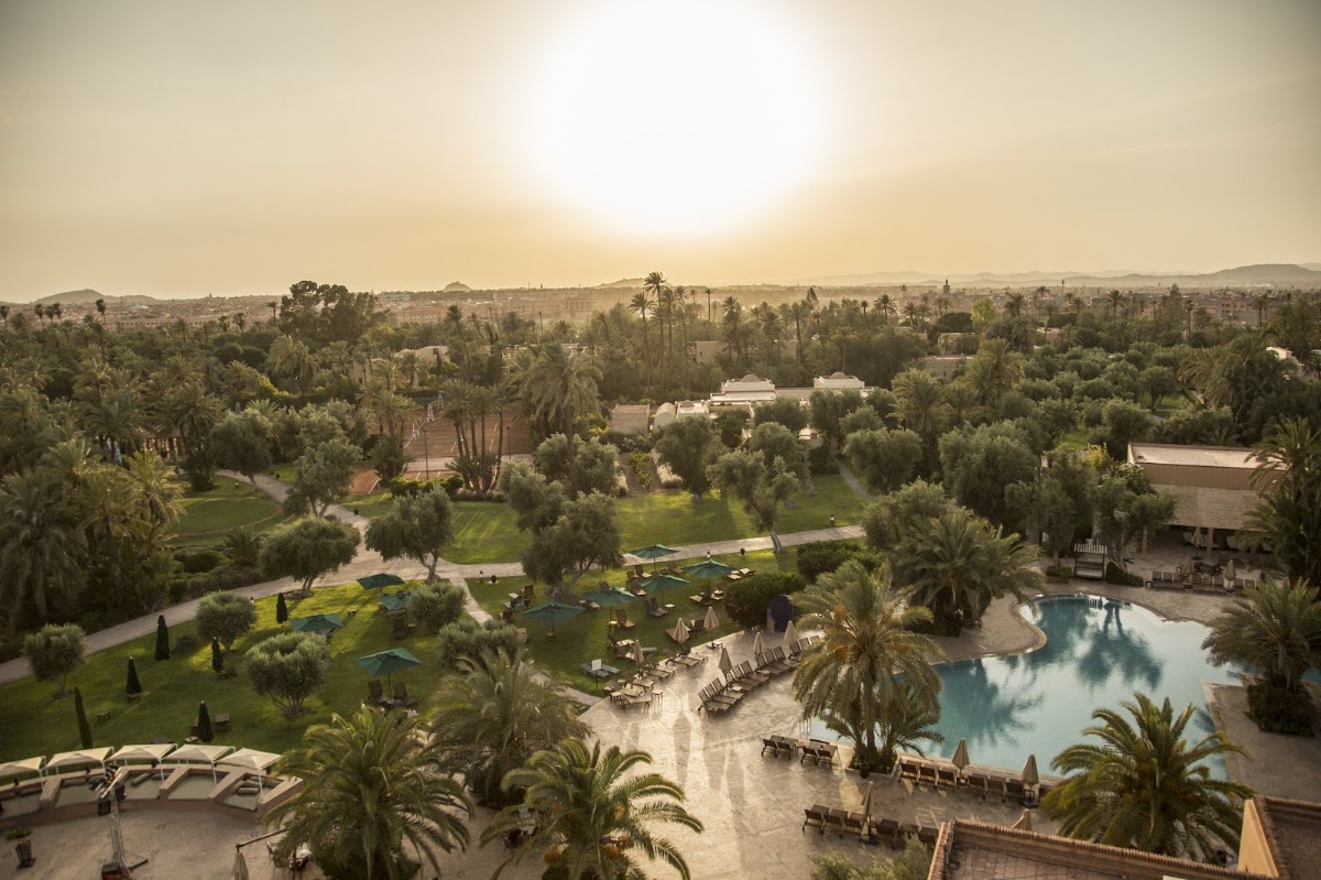 Aerial view of Club Med Marrakech La Palmeraie showing pool, loungers, palm-lined grounds, and Atlas Mountains in the distance at sunset.
