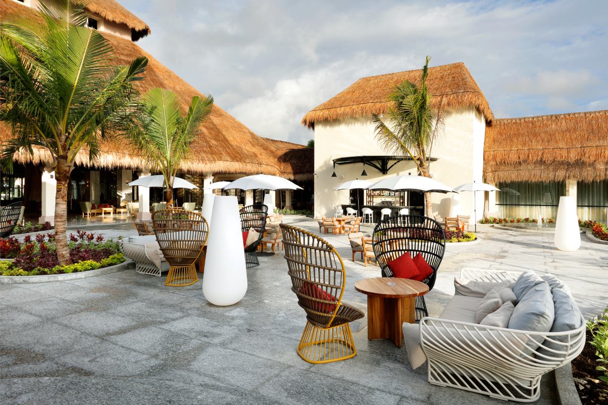 Resort courtyard with thatched-roof bungalows, woven chairs, white umbrellas, and lounges arranged around palm trees and flowering planters.