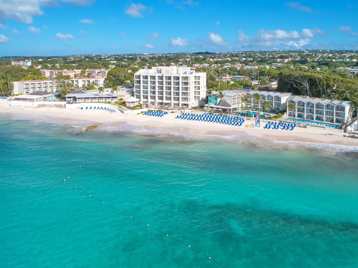 Aerial view of Sea Breeze Beach House resort on a sandy beach with turquoise water, blue loungers, and residential buildings beyond.