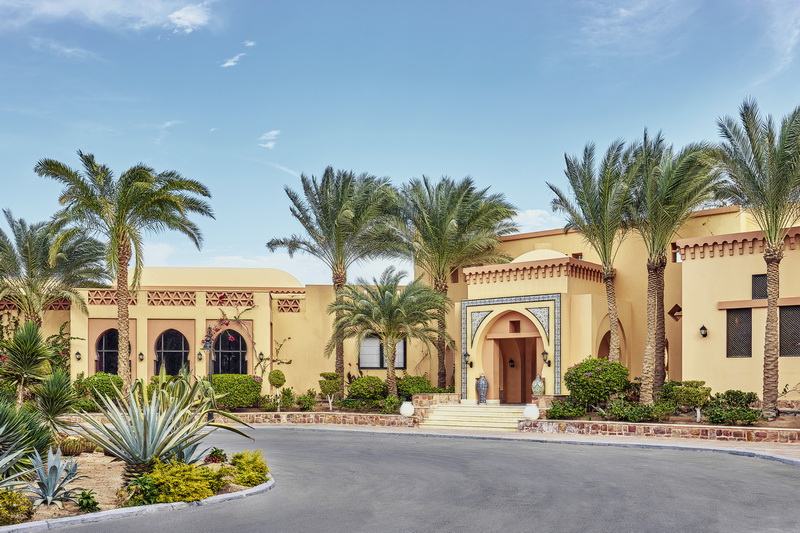 Cream-colored Moroccan-style resort buildings with arched doorways, palm trees, and desert landscaping under clear blue sky.
