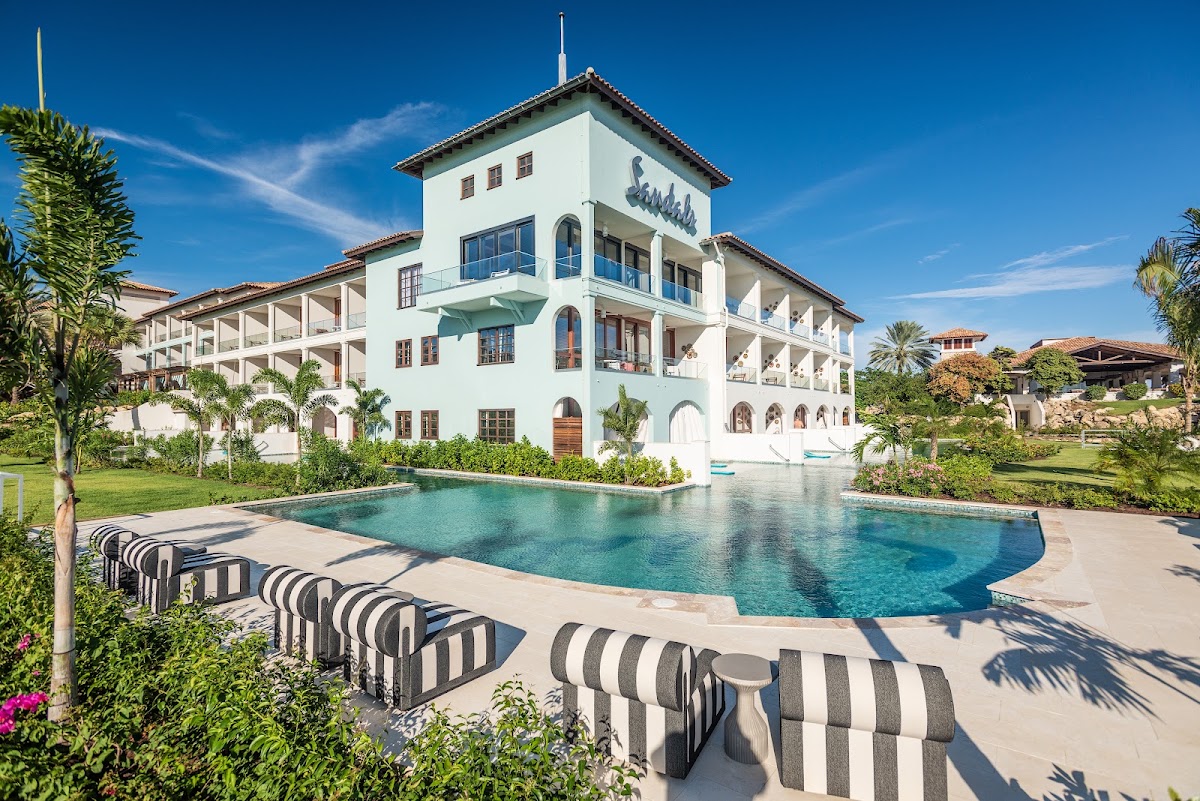 Striped lounge chairs line the pool deck at Sandals Royal Curaçao, a white colonial-style building with manicured grounds under clear blue sky.
