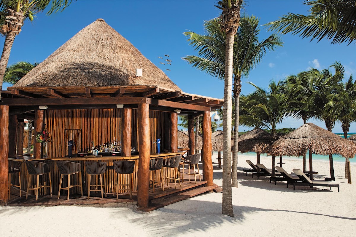 Thatched-roof beach bar with wooden structure and bar seating at Secrets Akumal Riviera Maya, with palm trees and lounge chairs on white sand.
