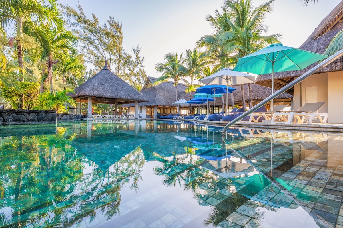Turquoise lagoon pool with thatched-roof bungalows, blue and green umbrellas, lounge chairs, and palm trees at Lagoon Attitude Hotel.