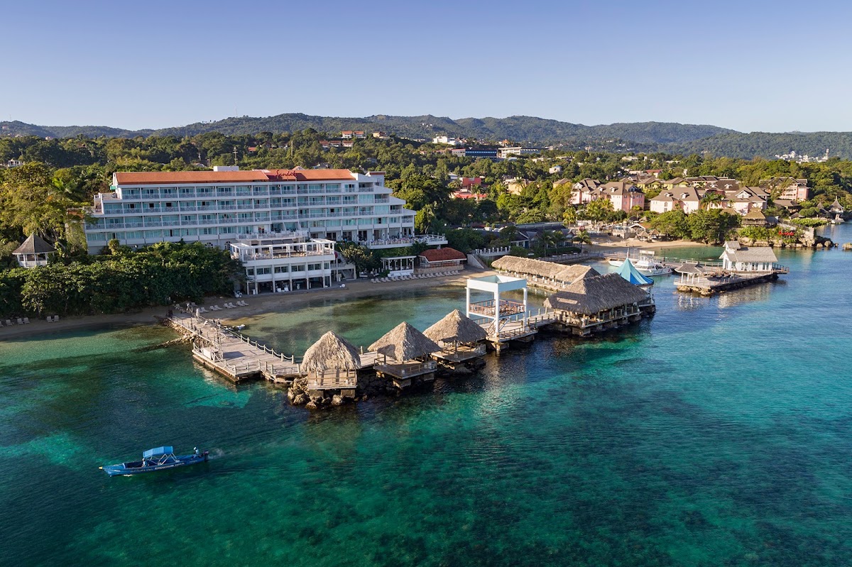 Blue and white multi-story resort building with thatched-roof overwater structures and beach access along turquoise Caribbean waters in Jamaica.