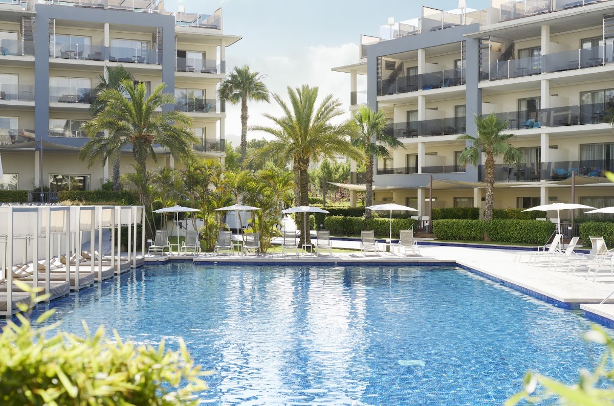 Blue rectangular pool with white umbrellas and loungers, surrounded by modern white and blue resort buildings and palm trees.