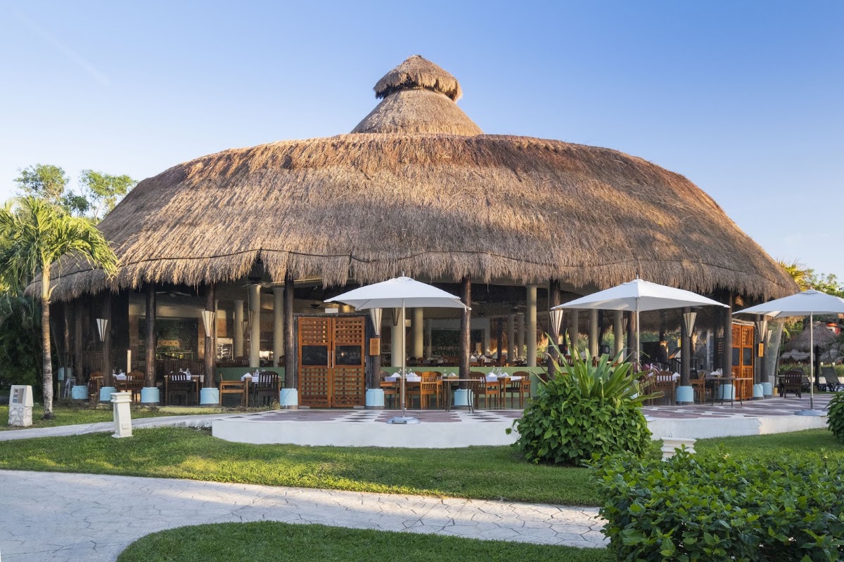 Large thatched-roof dining pavilion with white umbrellas, wooden doors, and palm trees at Iberostar Waves Cozumel.