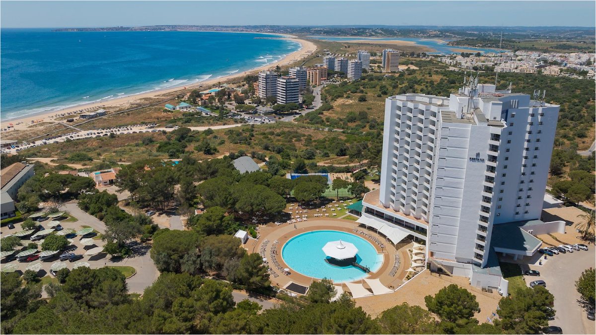 Pestana Blue Alvor Beach grounds with small pool, children's play area, and multicolored water slide, white loungers on grass beneath pine trees.