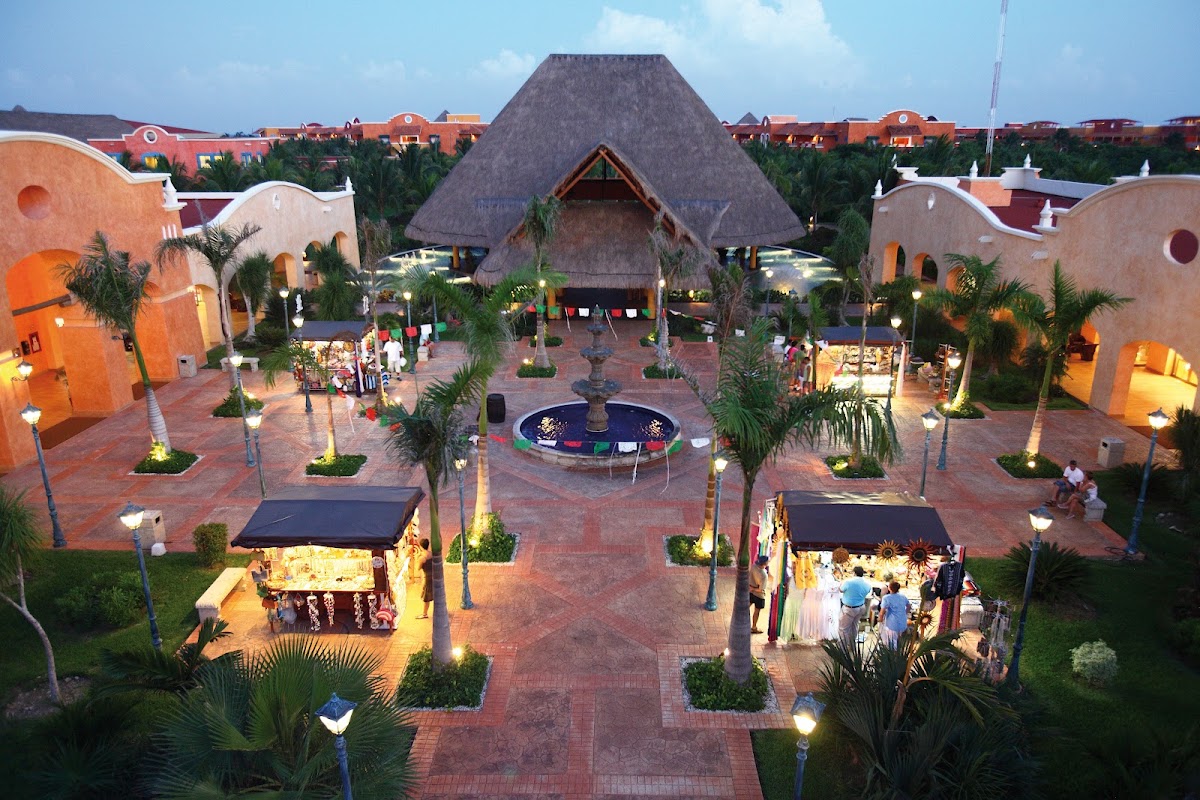 Central courtyard at dusk with thatched-roof pavilion, fountain, palm trees, lit shops, and orange colonial-style buildings surrounding the plaza.