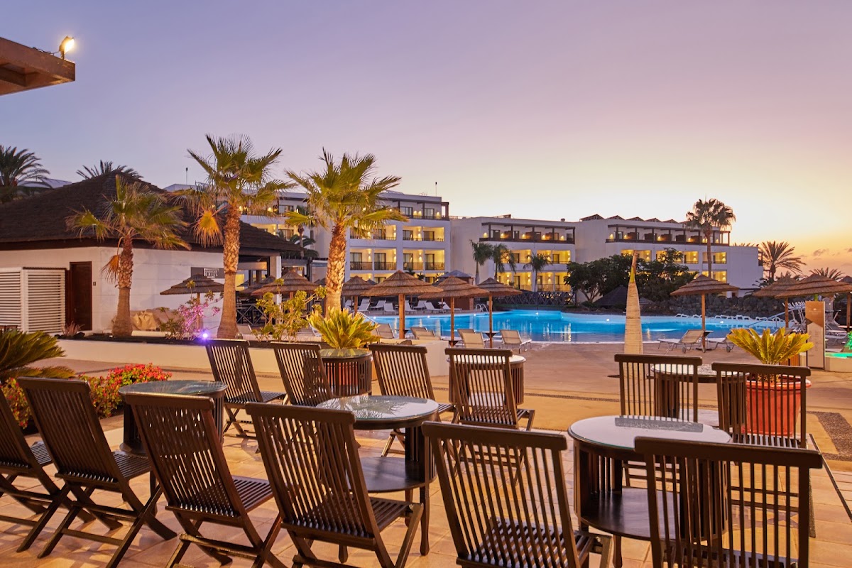 Lounge chairs and tables on a terrace overlooking the illuminated pool and white resort buildings at dusk, with palm trees and dark volcanic hills beyond.