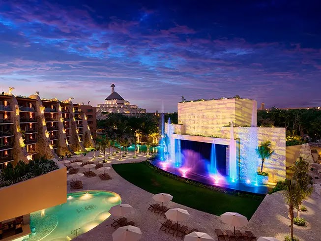 Nighttime resort courtyard with illuminated waterfall feature, pool, palm trees, and multi-story hotel buildings at dusk.