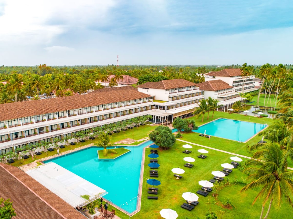 Resort buildings with terracotta roofs surround two turquoise pools and manicured lawns dotted with lounge chairs and umbrellas, palm trees throughout the grounds.
