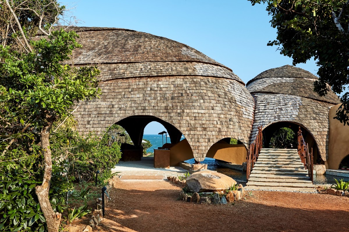 Blue slate-roofed dome structure at Wild Coast Tented Lodge with lit bar, wooden deck, and reflecting pool at dusk.