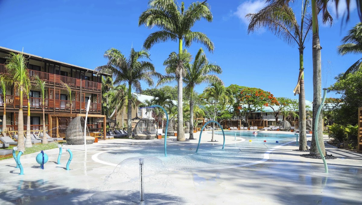 Shallow pool with blue water features and palm trees, wooden lodge buildings, and flowering trees under clear blue sky at Club Med La Pointe aux Canonniers.
