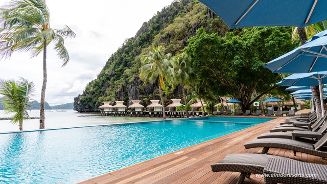 Turquoise pool with wooden deck and loungers overlooks limestone cliffs and traditional bungalows at El Nido Resorts Miniloc Island.