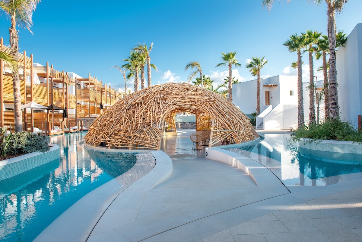 Woven wooden pergola spans between two pools at Stella Island Luxury Resort, with palm trees and white architecture surrounding the turquoise water.