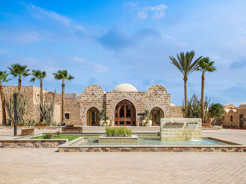 Stone building with domed roof and arched entrance at Movenpick Resort El Quseir, surrounded by palm trees and a reflection pool.