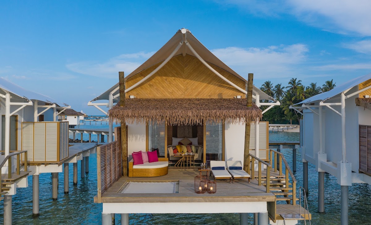 Overwater bungalow with thatched roof at Cinnamon Hakuraa Huraa Maldives, featuring a furnished deck with loungers and colorful cushioned seating.