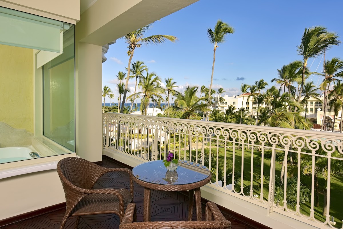 Wicker chairs and a small table on a balcony overlook palm trees, manicured grounds, and the ocean at JOIA Bávaro by Iberostar.
