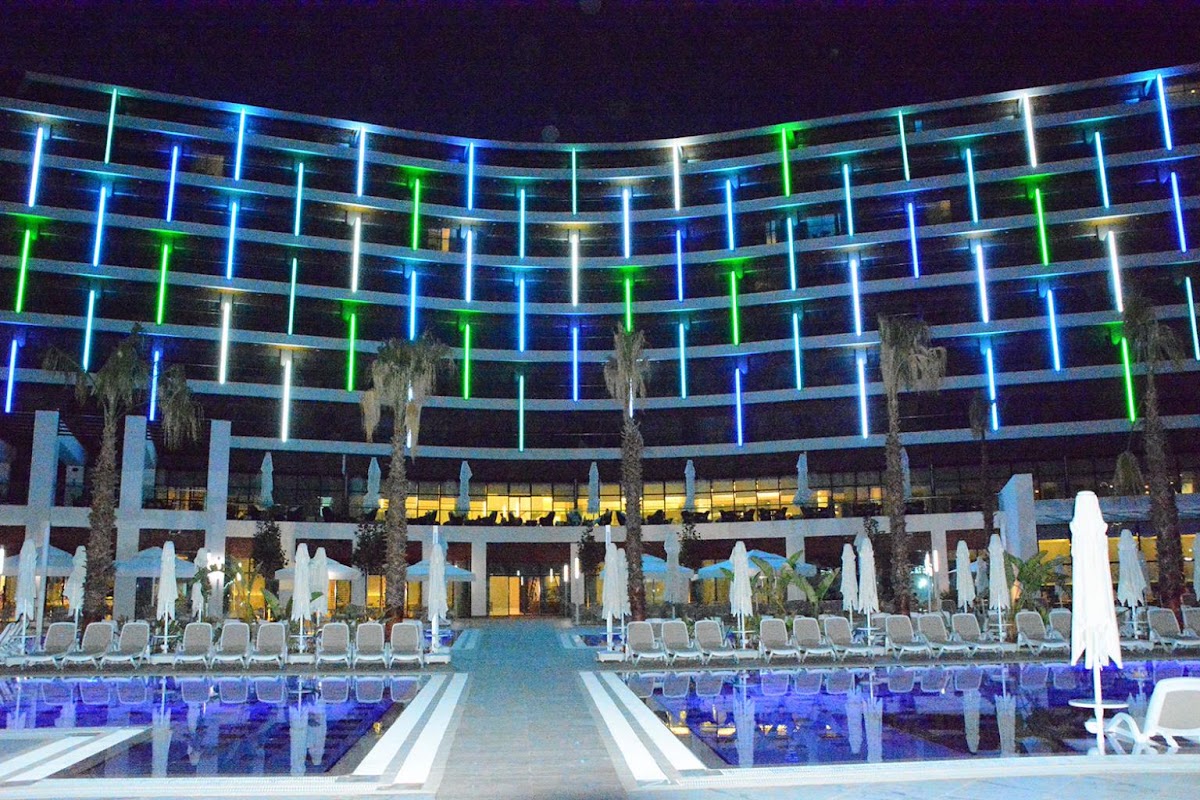 Night view of Wind of Lara Hotel & Spa's curved building facade with blue and green neon lights, pool deck with lounge chairs below, and palm trees.