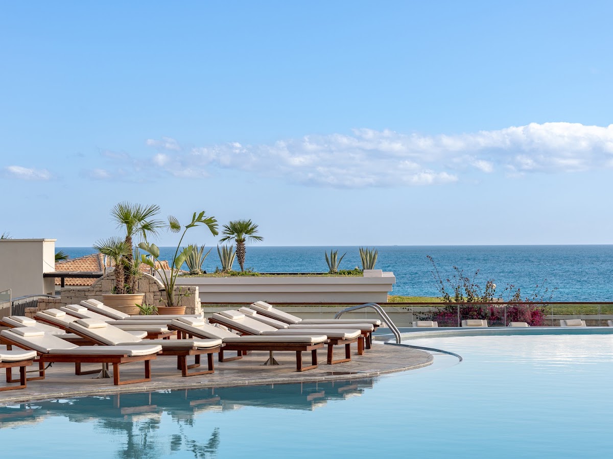 Loungers with white cushions line a curved pool overlooking the Mediterranean Sea at Grecotel Marine Palace & Aqua Park.