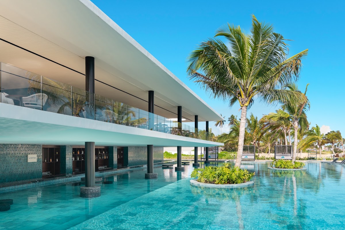 Modern resort building with turquoise pool, palm trees, and ocean view under clear blue sky at Live Aqua Beach Resort Punta Cana.