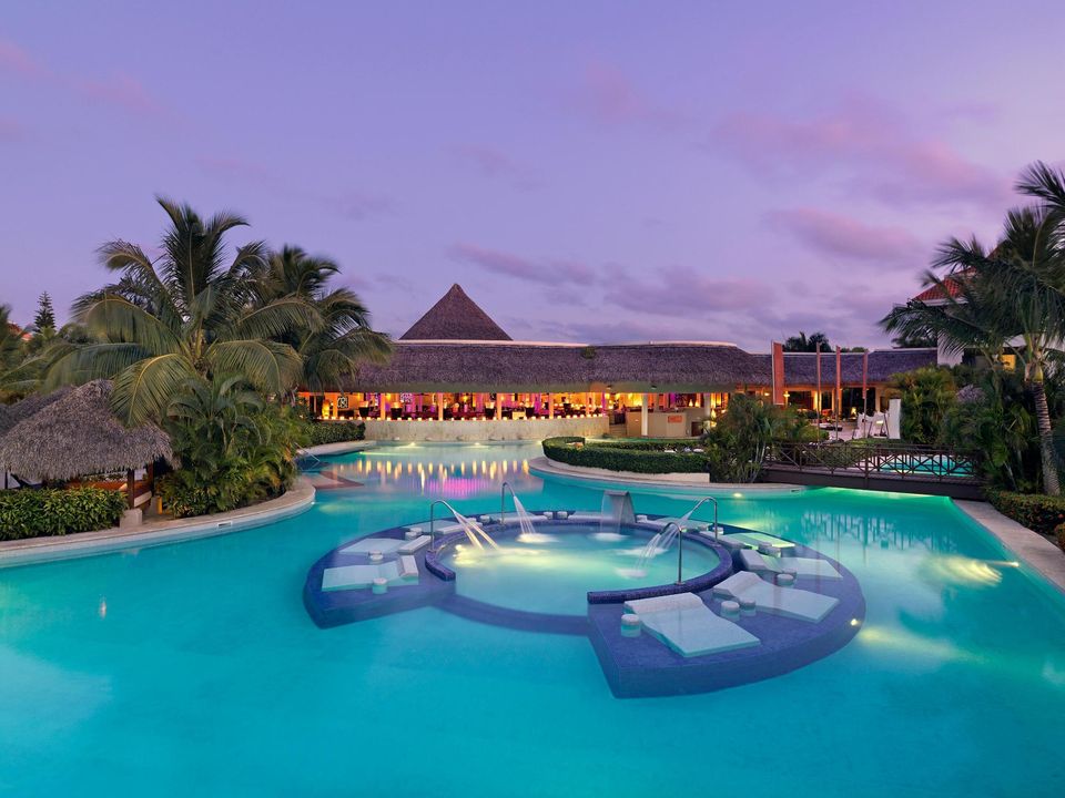 Turquoise lagoon pool with lit swim-up bar platforms at dusk, palm trees flanking an illuminated thatched-roof resort building beyond.