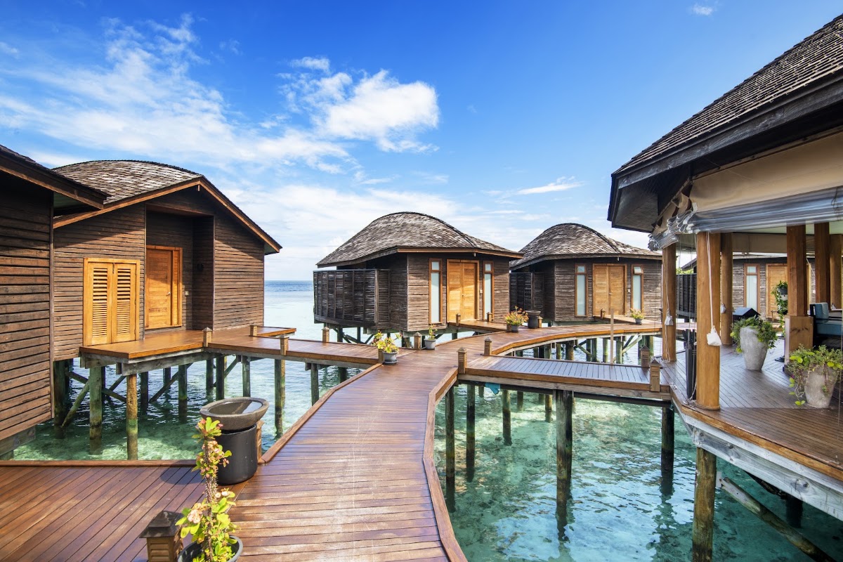 Wooden overwater bungalows with dark peaked roofs connected by a teak deck at Lily Beach Resort & Spa, with turquoise lagoon water and ocean horizon visible.