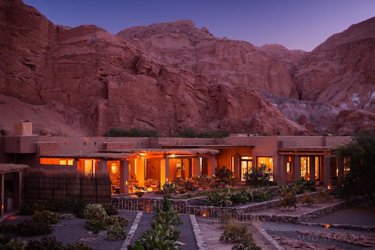 Desert resort buildings glow at dusk beneath towering red rock cliffs, with cacti and desert plants in the courtyard.