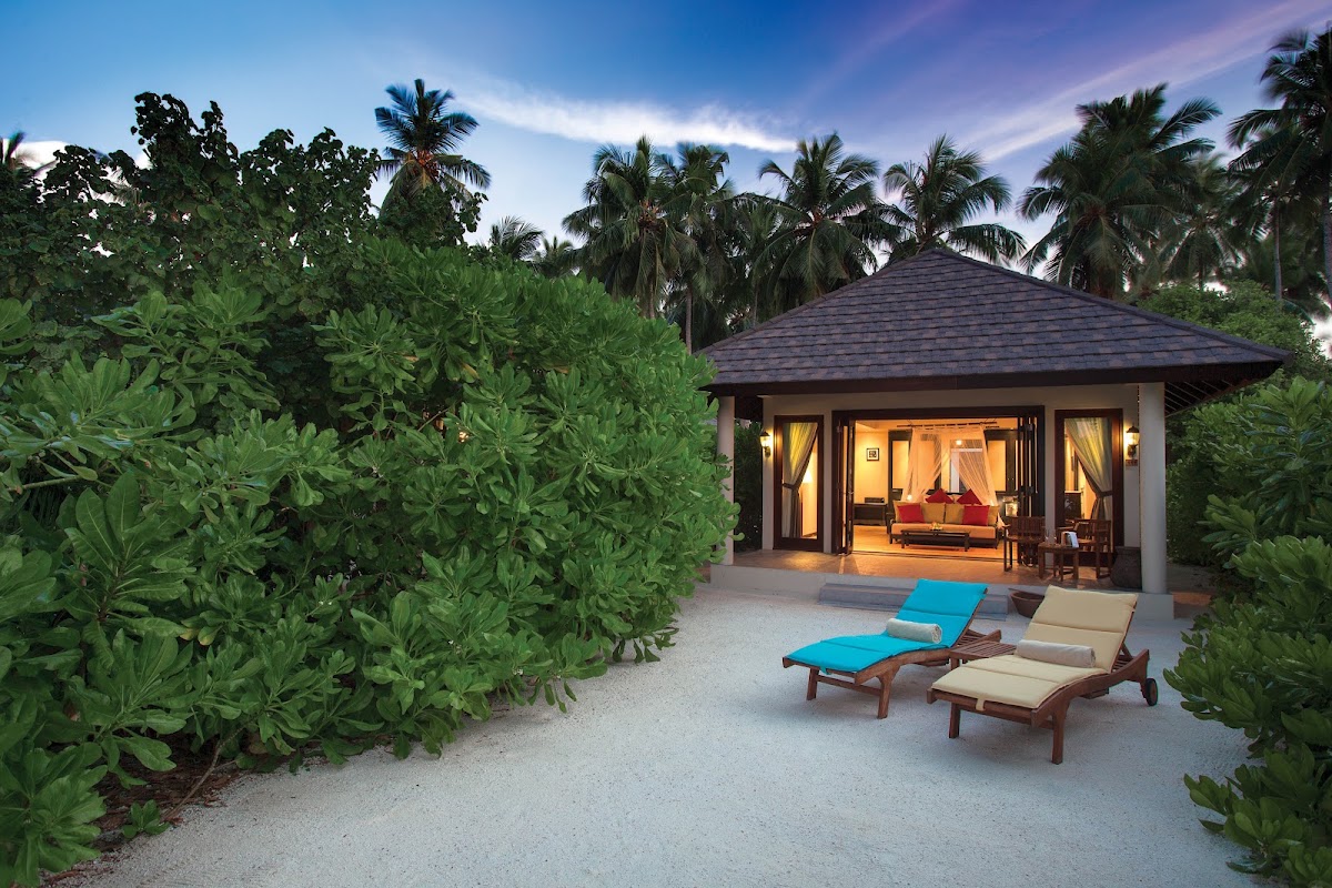 Lit villa with dark roof and lounge chairs on white sand, surrounded by green shrubs and palm trees at dusk.