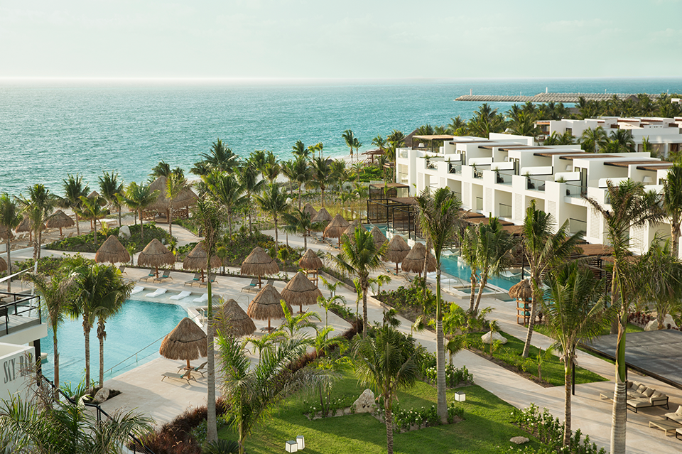 White modern buildings overlook palm-lined grounds with multiple pools and thatched palapas at Finest Playa Mujeres, facing turquoise Caribbean water.