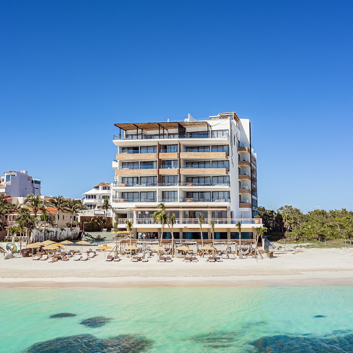 Modern beachfront resort with white and tan facade, ground-level lounge chairs and umbrellas on sand, turquoise shallow water in foreground, palm trees and clear blue sky.