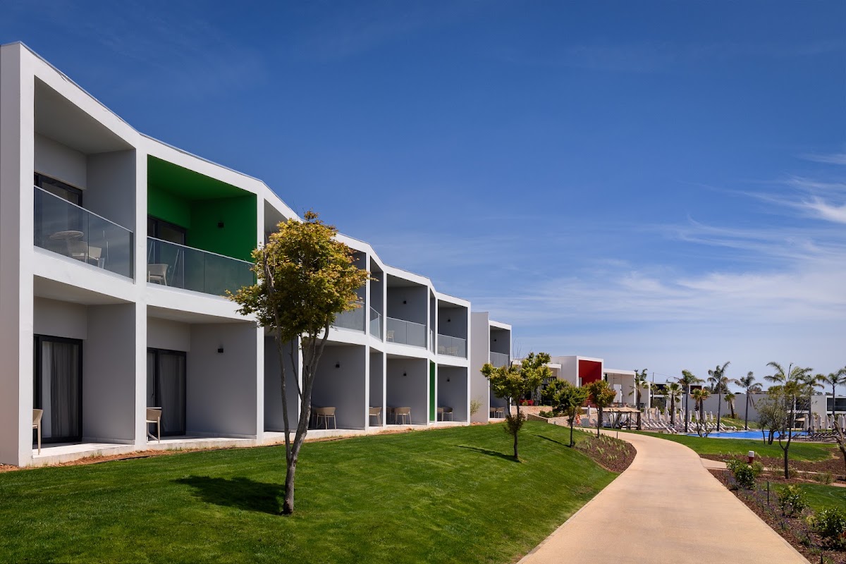 Modern white resort buildings with green accent panels line a manicured lawn and pathway at Tivoli Alvor Algarve, with palm trees and pool visible beyond.