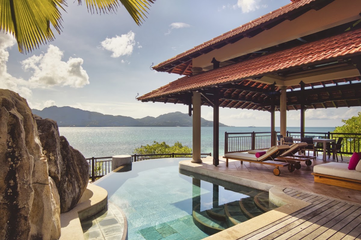 Oceanfront pavilion with terracotta roof overlooks turquoise infinity pool, loungers, and Seychelles mountains across the bay.