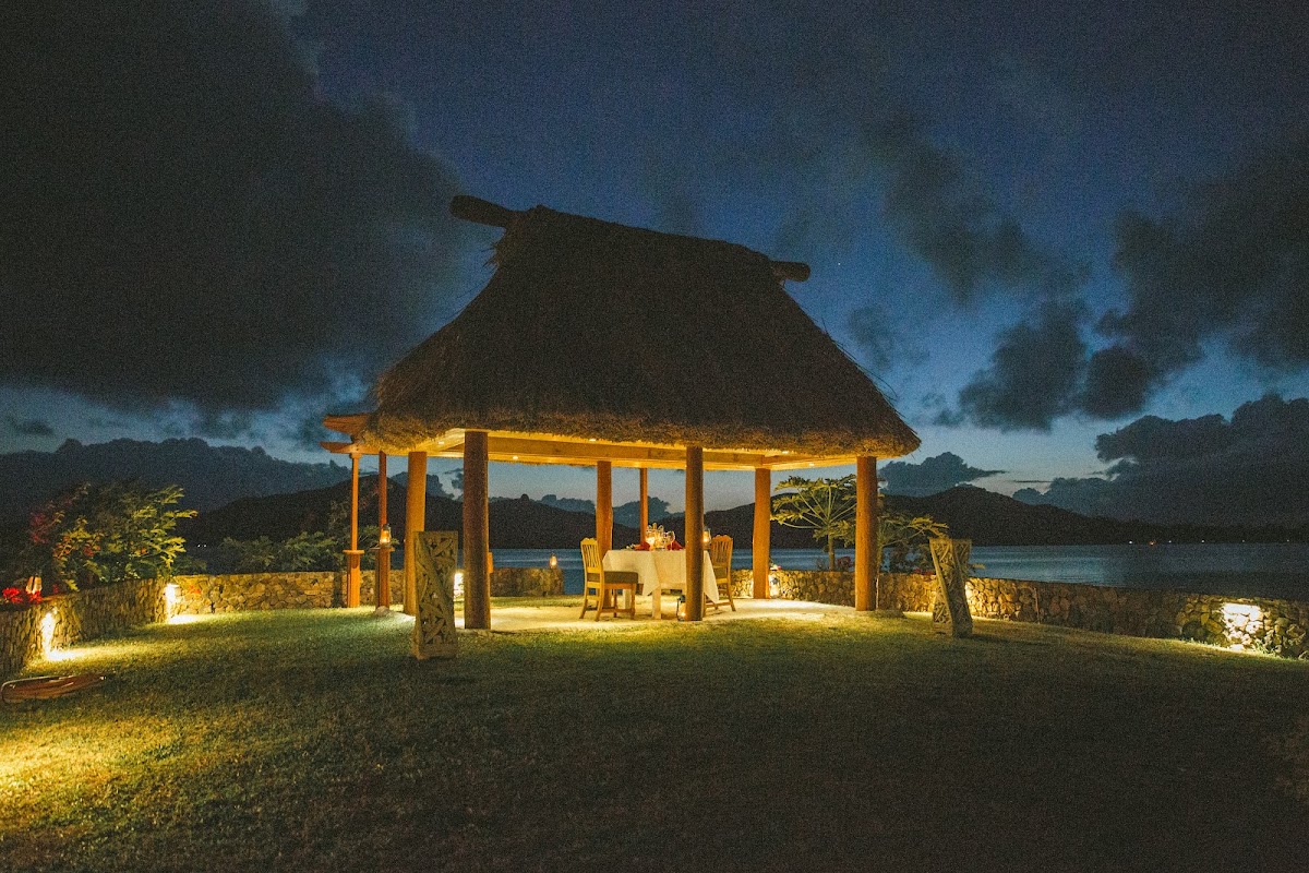 Illuminated thatched-roof pavilion with wooden posts overlooks a lagoon at dusk, surrounded by tropical vegetation and lit stone walls.