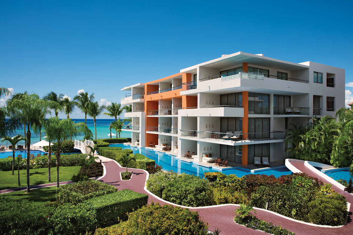 Modern white and orange resort building with curved balconies overlooks turquoise pool and ocean beyond, surrounded by palm trees and manicured landscaping.