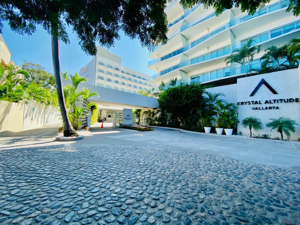 Krystal Altitude Vallarta's entrance courtyard with white modern buildings, palm trees, and patterned pavement under clear blue sky.