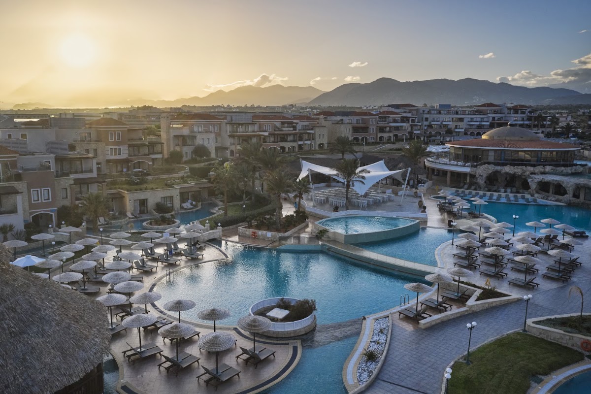 Resort pool complex with loungers and umbrellas overlooking Cretan buildings and mountains at sunset.