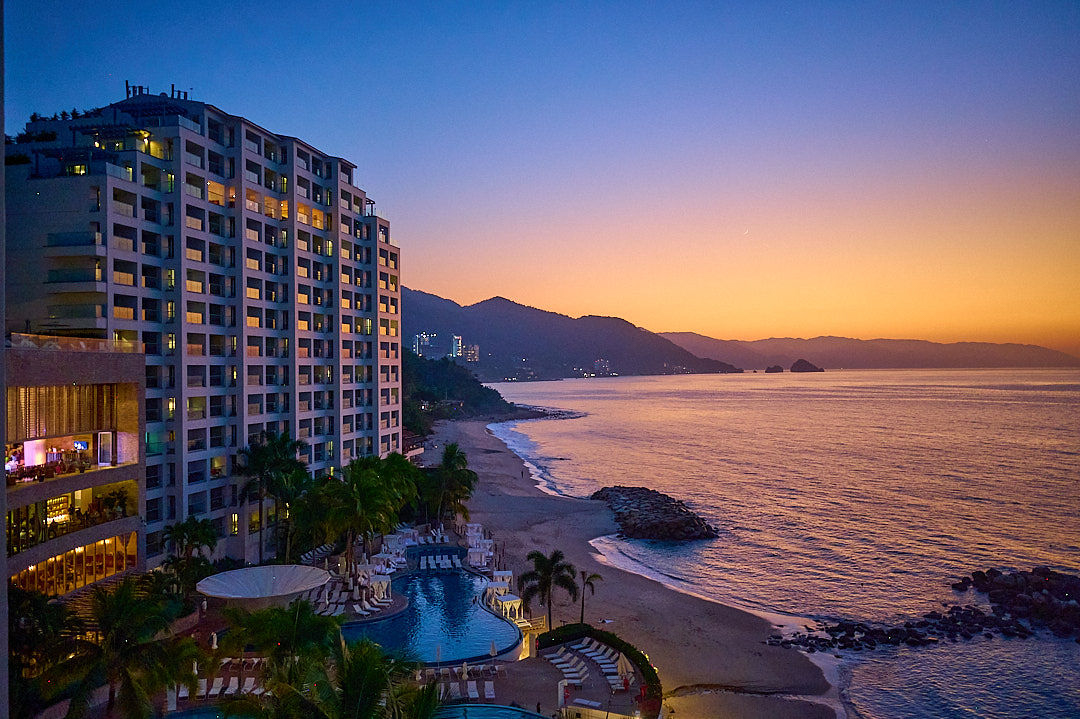 Hilton Vallarta Riviera beachfront with multi-story hotel towers, blue pools with red umbrellas, palm trees, and sandy beach with ocean waves and forested mountains beyond.