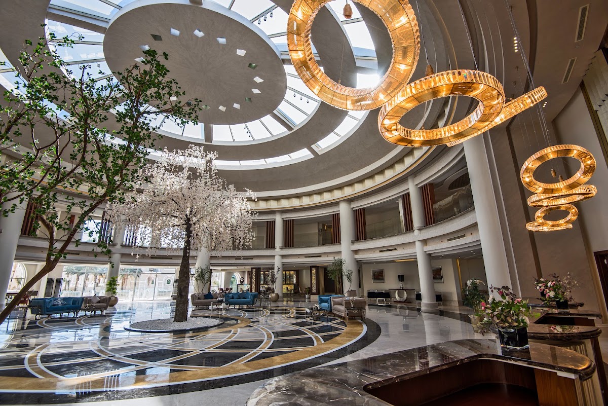 Modern atrium lobby with curved geometric ceiling, skylight, illuminated gold sculptural elements, polished marble floors, potted plants, and blue seating areas.