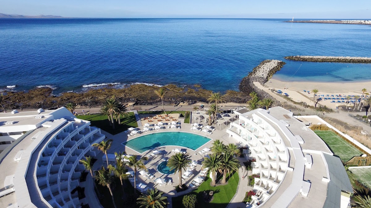 Aerial view of Iberostar Selection Lanzarote Park with curved white buildings, pool, palm trees, rocky coastline, and calm blue ocean.