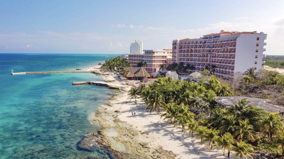 El Cozumeleño Beach Resort multi-story buildings overlook a sandy beach with palm trees and turquoise Caribbean waters.