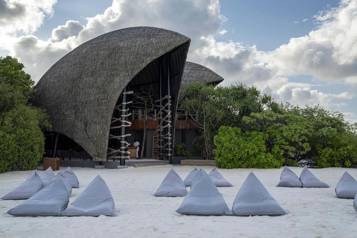 White bean bag loungers scatter across sand in front of a curved thatch-roof structure with dark wooden doors at NIVA Dhigali Maldives, surrounded by tropical vegetation.
